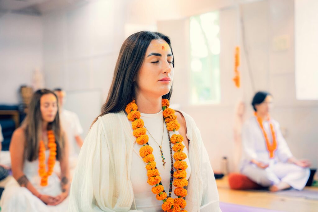 A group of women meditating in a tranquil indoor environment, wearing traditional clothing and flower garlands.