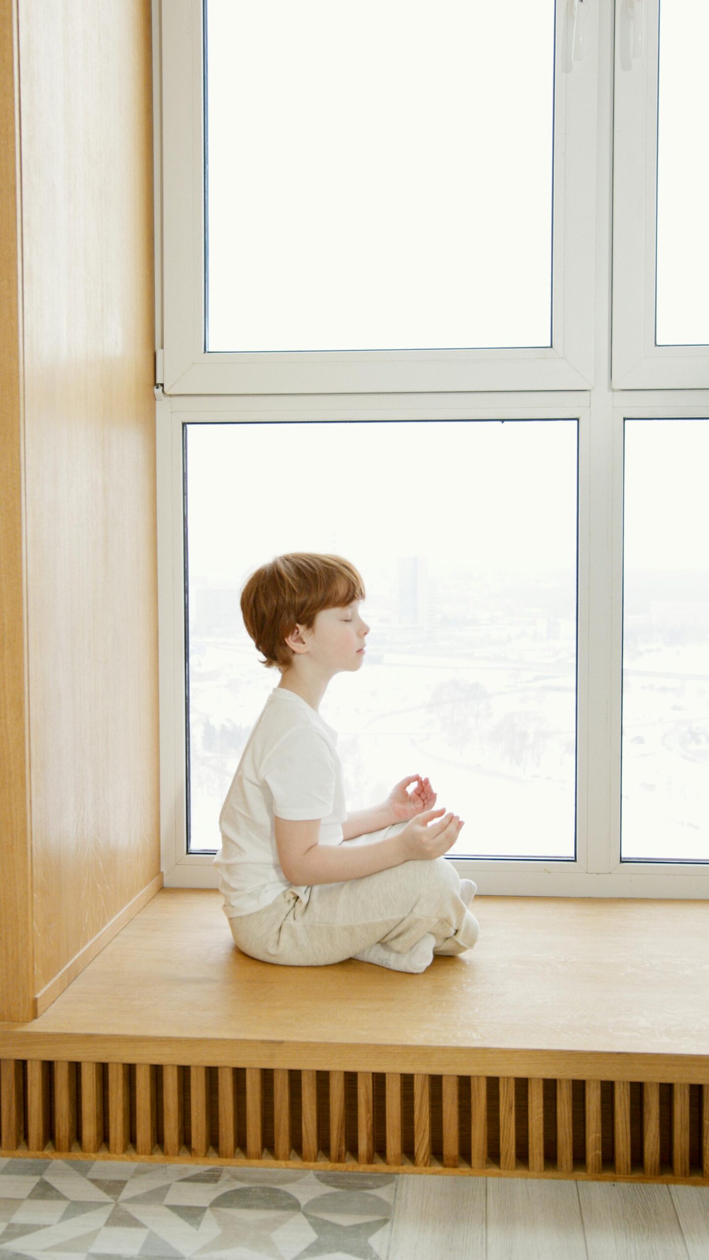 A young boy practices meditation indoors by a large, bright window, promoting peacefulness and focus.