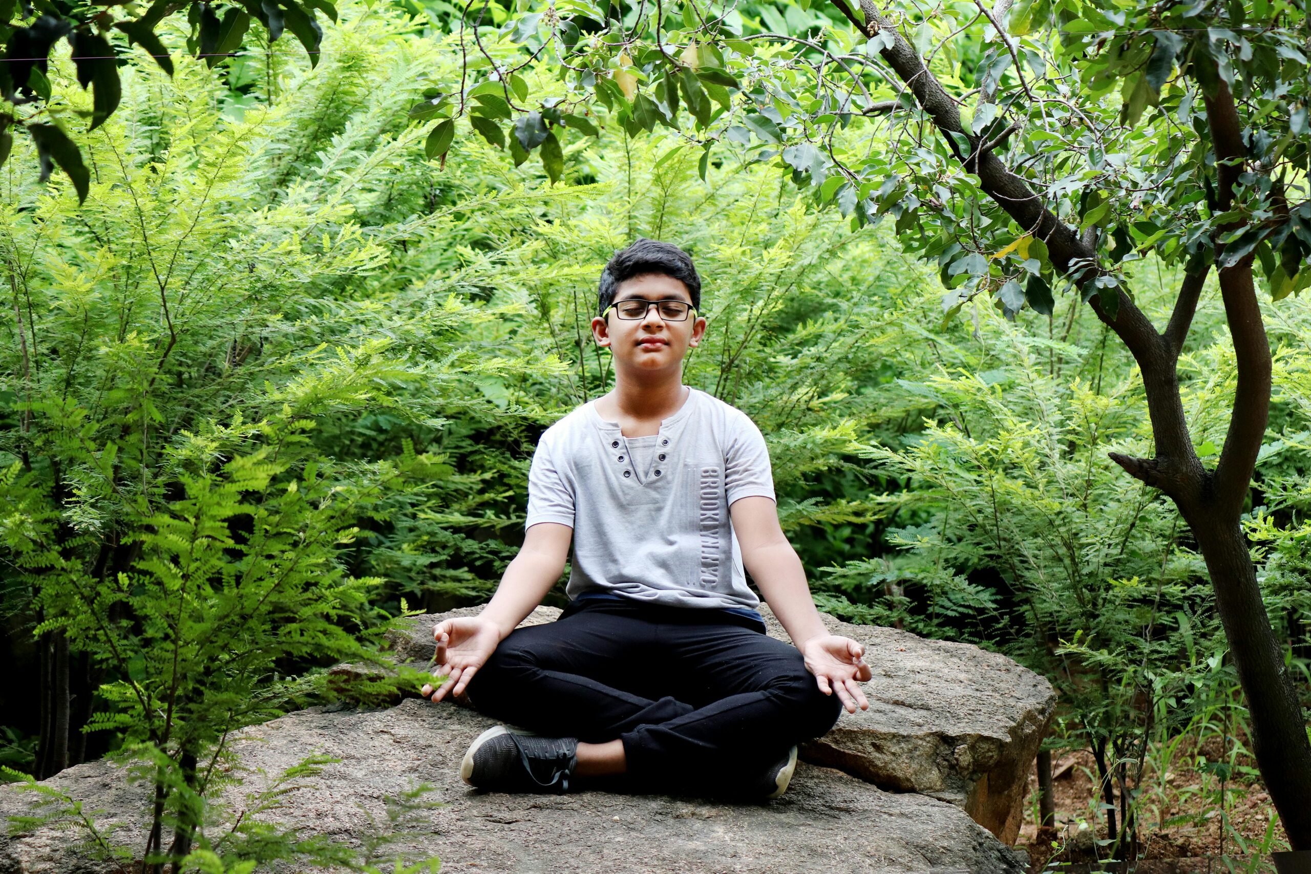 A teenager sitting in lotus pose meditating on a rock in a vibrant forest in Hyderabad, India.